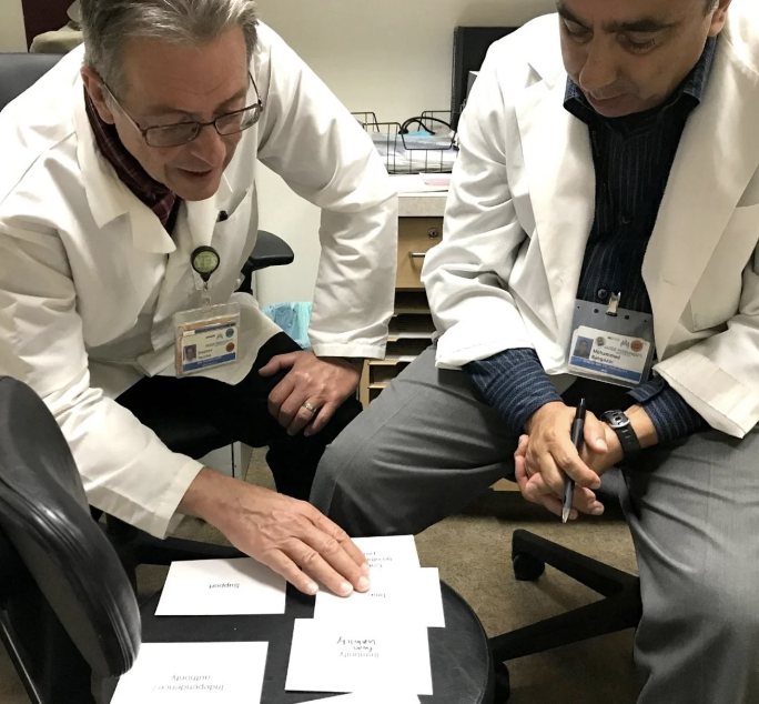 Two clinicians in white coats sit at a table, sorting and ranking small printed cards as part of a psychological-needs card sort activity.